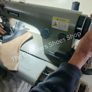May include: A close-up of a Brother industrial sewing machine in use. The machine is olive green and is stitching together a shoe component. The text "The Couplers Shoes Shop" is visible on the surface.