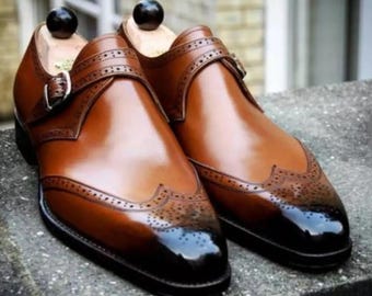 May include: A pair of brown leather shoes with a black toe cap and a single buckle strap. The shoes have a wingtip design and are made of a polished leather.