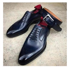 May include: A pair of navy blue leather dress shoes with intricate detailing on the toe, paired with a matching navy blue leather belt with a silver buckle. The shoes have laces and are placed on a neutral surface.