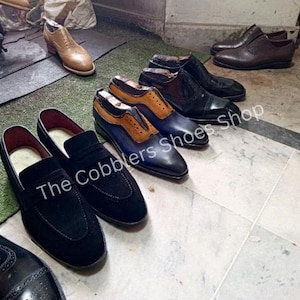 May include: A collection of handcrafted leather shoes in various styles and colors. The image features several pairs of dress shoes, including loafers and oxfords, in shades of black, brown, and blue. The Cobblers Shoes Shop is visible in the image.