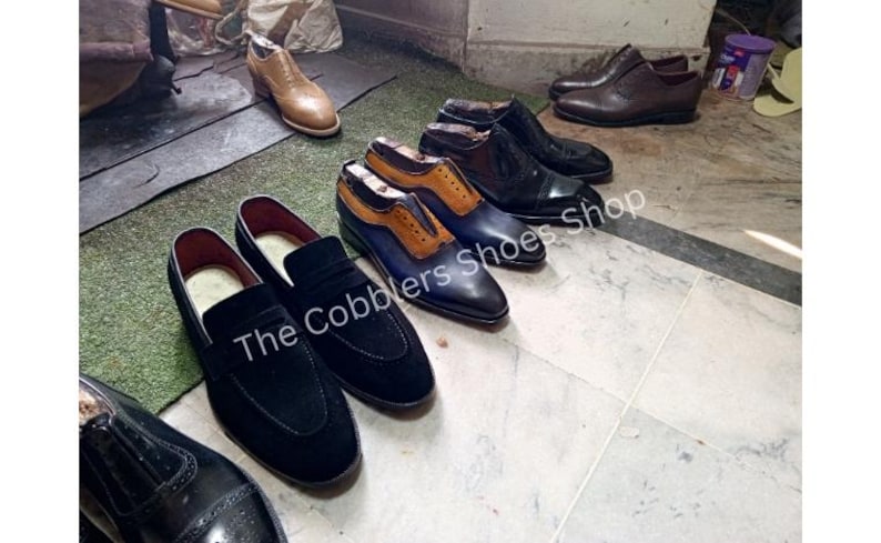 May include: A collection of handcrafted leather shoes in various styles and colors. The image features several pairs of dress shoes, including loafers and oxfords, in shades of black, brown, and blue. The Cobblers Shoes Shop is visible in the image.