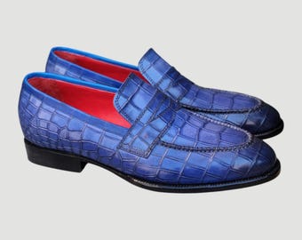 May include: A pair of blue leather loafers with a crocodile skin texture. The shoes have a red interior, black soles, and a classic slip-on design. The shoes are a vibrant shade of blue.