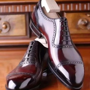 May include: A pair of polished, burgundy leather dress shoes with black soles and laces. The shoes feature decorative perforations along the seams and a wooden shoe tree inside. The shoes are displayed on a wooden surface.