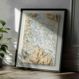 May include: A framed topographic map print featuring detailed terrain contours, elevation shading, and blue water bodies. The map displays geographical labels and grid lines in a neutral color palette, encased in a simple black frame.