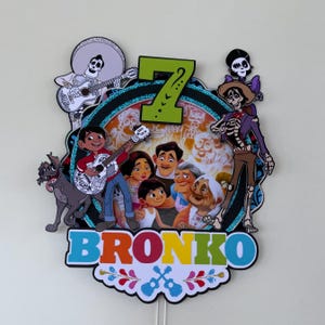 May include: A colorful cake topper featuring characters from the movie Coco. The design includes the number 7, the name "BRONKO", and images of guitars and skeletons. The characters are set against a background of skulls.