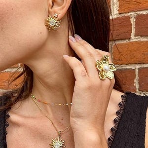 Peut inclure: Ensemble de bijoux dorés comprenant un collier pendentif soleil, des boucles d'oreilles assorties et une bague fleur. Les boucles d'oreilles et le pendentif ont un groupe central de petites pierres claires. La bague a un centre en forme de perle.