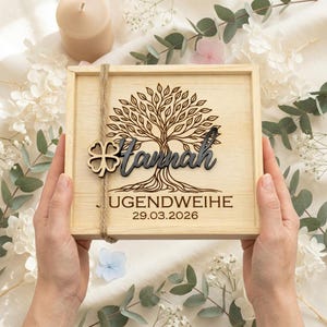 Könnte beinhalten: Eine helle Holzkiste mit einem Baumdesign, dem Namen "Hannah" und dem Text "JUGENDWEIHE 29.03.2026". Die Kiste ist mit Bindfaden gebunden und hat einen vierblättrigen Kleeblatt-Anhänger. Die Kiste wird von zwei Händen gehalten.