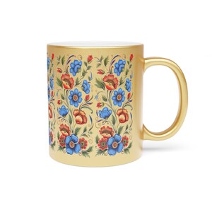 Puede incluir: Una taza de cerámica dorada con un patrón floral en tonos azules, rojos y verdes. La taza tiene un interior blanco y una asa redondeada.
