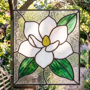 Op de afbeelding: Een vierkant glas-in-lood paneel met een centrale witte magnolia bloem met een geel hart en groene bladeren. De glasstukken worden bij elkaar gehouden door donkere metalen profielen en het geheel hangt aan een dunne zilveren metalen ketting.