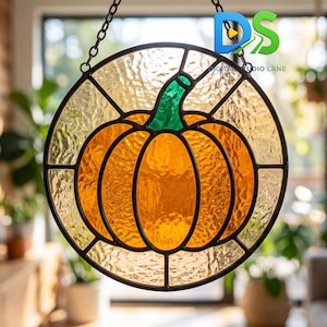 May include: A circular stained glass suncatcher featuring an orange pumpkin with a green stem, encased in a black metal frame. The glass has a textured, rippled finish. It hangs from a black metal chain and includes a small logo reading DS.