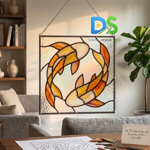 Könnte beinhalten: Ein Buntglasfenster mit zwei Koi-Karpfen in Orange und Gelb, die ein kreisförmiges Muster bilden. Das Kunstwerk ist in dunklem Metall gerahmt und an einer Kette aufgehängt. Das Fenster ist in einem hellen Raum ausgestellt.