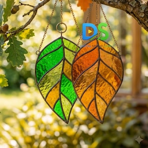 Può includere: Due ornamenti a forma di foglia in vetro colorato appesi a un ramo d'albero. Una foglia è di un verde vivace, l'altra è di un caldo arancione ambrato. Entrambe presentano linee di saldatura in metallo scuro che definiscono le venature.