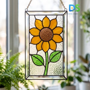 May include: A rectangular stained glass suncatcher featuring a central yellow sunflower with a textured copper-colored center and two green leaves. The piece is framed in dark metal and hangs from a gold-toned chain against a window background.