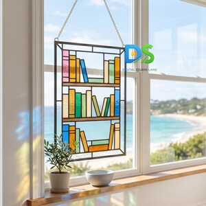May include: A rectangular stained glass suncatcher featuring a three-shelf bookcase design filled with multicolored glass books. The piece is framed in dark metal and hangs from a gold-toned chain, displaying various hues of blue, green, yellow, and pink.