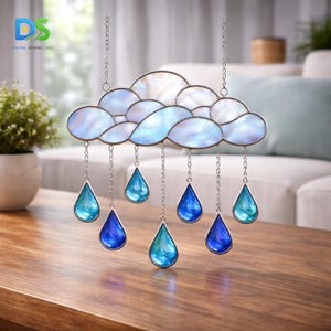 Puede incluir: Una nube de vidriera con gotas colgantes. La nube es de vidrio blanco y azul claro, con detalles de cadena plateada. Las gotas son en tonos azules y turquesas. Una pieza decorativa para ventana o pared.