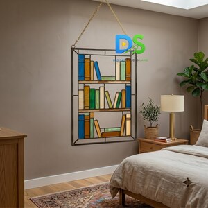 May include: A rectangular stained glass wall hanging featuring a three-shelf bookcase design with various colored glass books in blue, green, yellow, and brown tones. The piece is framed in dark metal and suspended by a gold-toned metal chain.