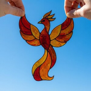 Può includere: Un acchiappasole in vetro colorato a forma di fenice con ali spiegate. Il pezzo è composto da singoli segmenti di vetro rosso, arancione e giallo tenuti insieme da linee di saldatura in metallo scuro, creando un vibrante effetto di fuoco.