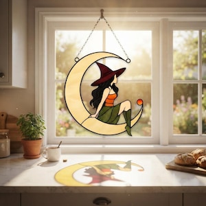 May include: A stained glass suncatcher shaped like a crescent moon with a figure wearing a dark red pointed hat, an orange top, and a green skirt sitting on the moon. The piece hangs from a silver chain and casts a colorful shadow on the counter.