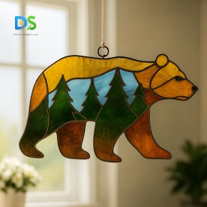 Puede incluir: Atrapasoles de oso de vidriera con una escena de bosque. El oso es marrón, naranja y amarillo, con un cielo azul y árboles verdes. El atrapasoles cuelga de un cordón beige y tiene un anillo de metal.