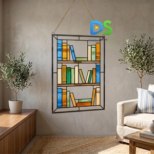 May include: A rectangular stained glass suncatcher featuring a three-shelf bookcase design with various colored glass books. The piece is suspended by a gold-toned metal chain and includes a dark metal frame outlining the shelves and books.