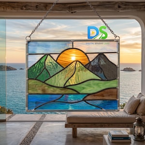 May include: A rectangular stained glass panel featuring a landscape scene with a bright orange sun rising behind green and dark gray mountains, set above a blue water foreground. The piece is framed in metal and suspended by two silver chains.