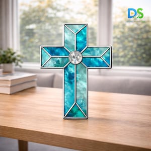 May include: A stained glass cross with a silver metal frame. The cross features shades of blue and teal glass with a clear, round jewel in the center. The cross is displayed on a wooden table.