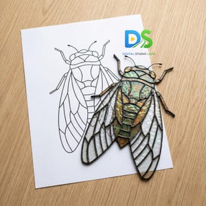 Puede incluir: Una cigarra de vidriera con alas iridiscentes, posada sobre un papel blanco con un contorno de cigarra. El insecto presenta tonos de verde, dorado y blanco. La imagen está sobre una superficie de madera.