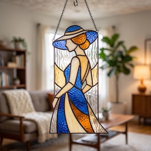 May include: A stained glass suncatcher featuring a profile of a figure wearing a wide-brimmed hat. The design uses textured blue, amber, and clear glass pieces held together by dark metal lead lines, suspended by a silver-coloured metal chain.