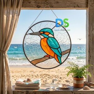 Puede incluir: Un atrapasueños circular de vidrieras que muestra un martín pescador con plumaje naranja brillante y azul cerceta posado en una rama. El fondo del panel de vidrio tiene una textura blanca agrietada. La pieza cuelga de una cadena de metal.