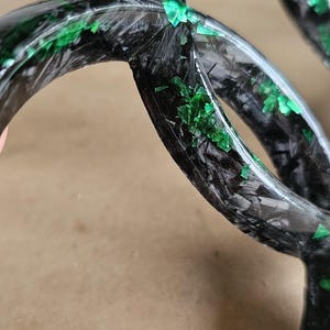 May include: Close-up of a black and green decorative object. The object has a curved, circular shape with a textured, carbon fiber appearance. Green accents are scattered across the surface.