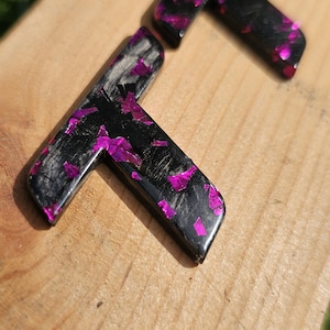 Pode incluir: Três letras F de resina preta com glitter rosa vibrante embutido, repousando sobre uma superfície de madeira clara. As letras estão anguladas, mostrando o brilho do glitter. O fundo está desfocado.