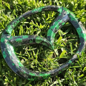 May include: A black and green car emblem with a glittery finish. The emblem is shaped like a circle with a split in the top. The emblem is sitting on a bed of green grass.