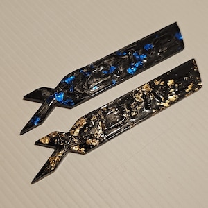 May include: Two black resin pieces with a unique shape, one with blue and the other with gold leaf accents. The pieces have a pointed end and a flared, wing-like design at the other end. They are displayed on a light-colored surface.