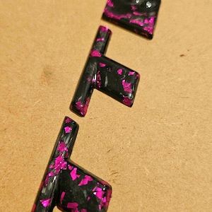 Pode incluir: Três chaveiros de resina preta com incrustações de glitter rosa são exibidos em uma superfície marrom clara. Os chaveiros têm o formato de uma chave e são divididos em três peças separadas.
