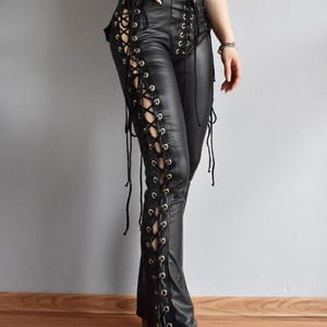 Könnte beinhalten: Schwarze Kunstlederhose mit hoher Taille, silberfarbenen Metallösen und schwarzer Schnürung, die sich über die gesamte Länge des äußeren Beins erstreckt. Die Hose hat eine eng anliegende Passform und eine glatte, matte Oberfläche.