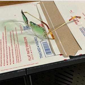 May include: Gold-framed sunglasses with green lenses, resting on a cardboard box. The box features the text "PRIORITY MAIL" and "FLAT RATE ENVELOPE". The glasses have a unique, angular design.