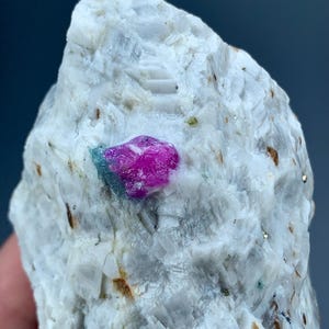 May include: A rough, white rock with a vibrant, fuchsia-colored crystal embedded within. The crystal has a slightly blue-green hue at its base. The rock's surface has a textured appearance with small, brown and gold flecks.