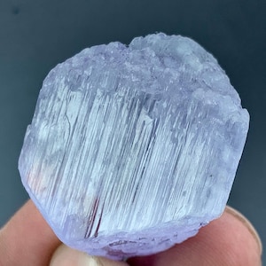 May include: A close-up of a light purple crystal with a rectangular shape. The crystal has a rough, textured surface with vertical striations. The color is a pale lavender, and the crystal appears to be translucent.