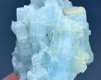 May include: A cluster of light blue aquamarine crystals. The crystals have a layered, columnar structure with a translucent appearance. Some areas show a slight yellow hue. The mineral is held against a dark background.