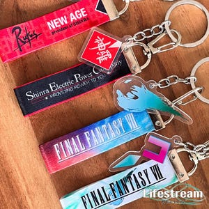 Könnte beinhalten: Schlüsselanhänger mit verschiedenen Designs und Texten. Einer hat den Text "NEW AGE president of SHINRA" auf einem roten Band. Ein anderer hat "Shinra Electric Power Co. PROVIDING POWER TO YOU" auf einem schwarzen Band. Weitere Schlüsselanhänger zeigen "FINAL FANTASY VII" und ein Logo.