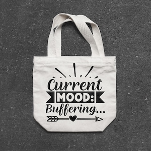 May include: A small, off-white canvas tote bag with a black graphic that reads "Current MOOD: Buffering...". The design includes an arrow, heart, and decorative lines. The bag has two short handles.