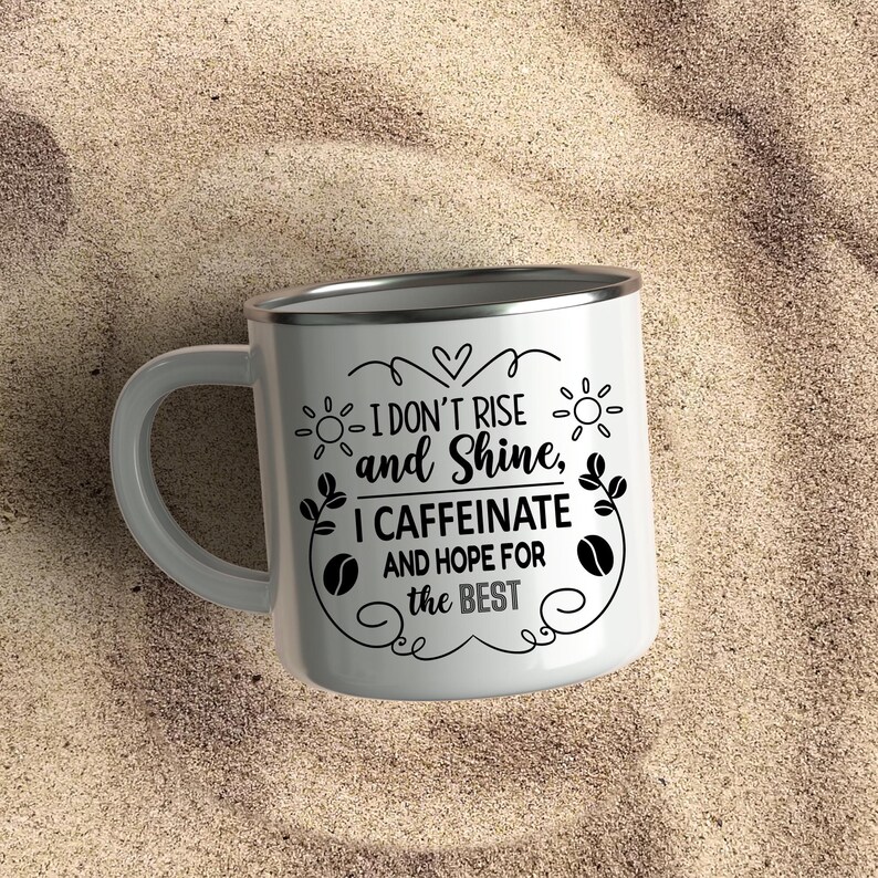 May include: White enamel coffee mug with a silver rim and handle. The mug features black text that reads "I don't rise and shine, I caffeinate and hope for the best." Decorative sun and coffee bean graphics surround the text. Perfect for coffee lovers.