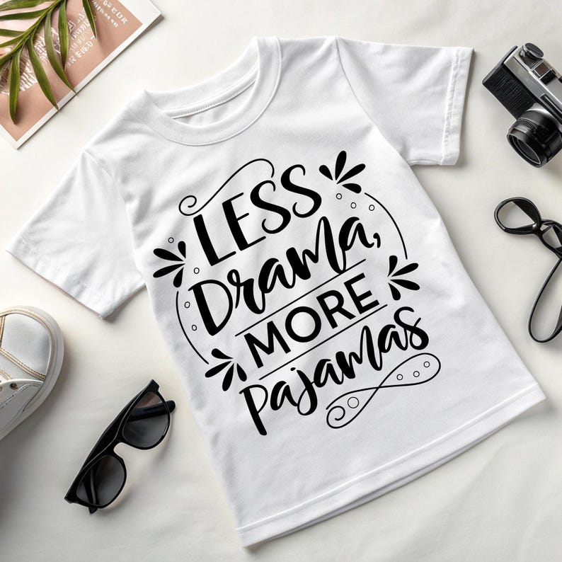 May include: White t-shirt with the text "LESS Drama, MORE Pajamas" in black script. The shirt is a crew neck style and is displayed on a white surface with sunglasses, a camera, and other accessories.