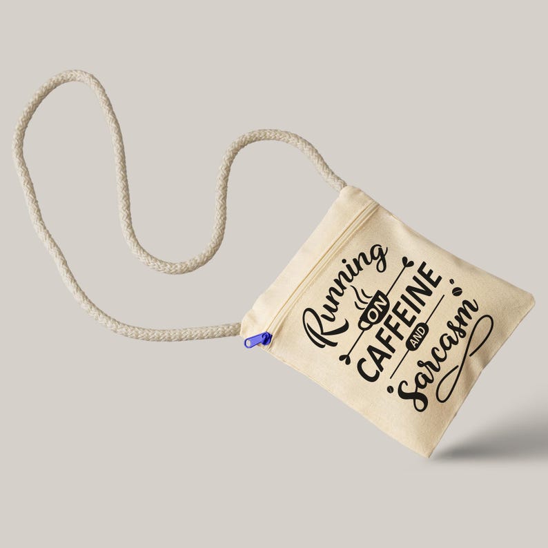 May include: A beige canvas crossbody bag with a cream-colored rope strap. The bag features a blue zipper and the text "Running on Caffeine and Sarcasm" in black script. The bag is designed to be worn over the shoulder.