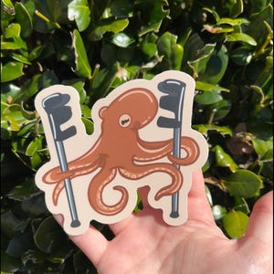 May include: A sticker of a brown octopus holding two black golf clubs. The octopus has eight tentacles and is smiling. The sticker is on a white background.