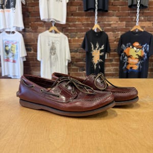 May include: A pair of dark brown leather boat shoes featuring a moccasin-style toe, rawhide laces threaded through metal eyelets along the sides, and a low-profile rubber sole. The Timberland logo is embossed on the side of the leather upper.