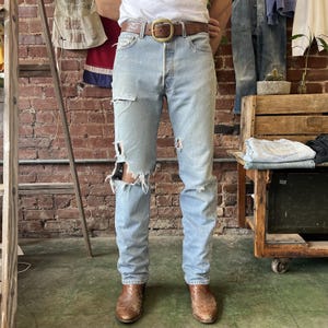 May include: Light blue denim jeans featuring a distressed finish with rips and frayed edges on the thighs and knees. The jeans are secured with a brown leather belt that has a large, rounded brass-toned metal buckle, paired with brown leather boots.