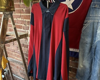 Puede incluir: Una camisa tipo polo de manga larga con un diseño de bloques de color llamativo con paneles verticales en azul marino intenso y rojo brillante. La prenda cuenta con un cuello clásico, una tapeta con botones y mangas largas.