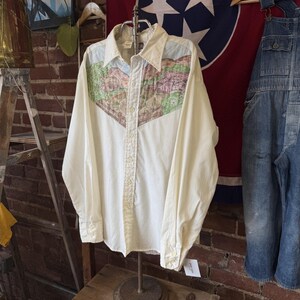 May include: A long-sleeve off-white button-down shirt featuring a large, V-shaped yoke panel across the chest and shoulders. The panel displays a detailed landscape illustration in shades of green, brown, and blue, depicting a rural scene.