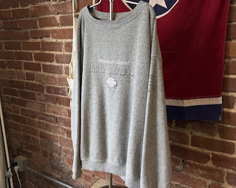 May include: A heather grey long-sleeve sweatshirt featuring a ribbed crew neckline and cuffs. The front displays the text American Legend and Harley-Davidson in dark lettering, accompanied by a small shield-shaped logo graphic.